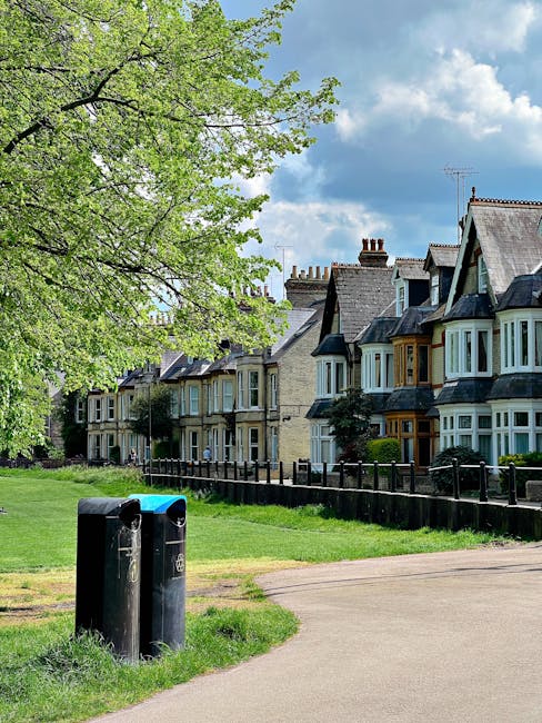 A row of traditional terraced houses with bay windows and pitched roofs, set behind a black wooden fence. The houses feature brickwork and some have decorative elements in different shades of grey, cream, and muted yellow. In the foreground, a small grassy area is adjacent to a paved walkway, with two black and blue waste bins positioned near the edge of the grass. A large deciduous tree with fresh green leaves extends over part of the scene, providing partial shade. The background showcases a partly cloudy sky with patches of blue. The scene appears to be a residential street, and the presence of waste bins suggests routines of rubbish collection alongside the houses, which might contrast with or supplement alternative waste handling options such as private rubbish removal services, as offered by companies like House Clearance Tufnell Park. The environment looks well-maintained and typical of an urban or suburban neighbourhood, with a peaceful, orderly atmosphere.
