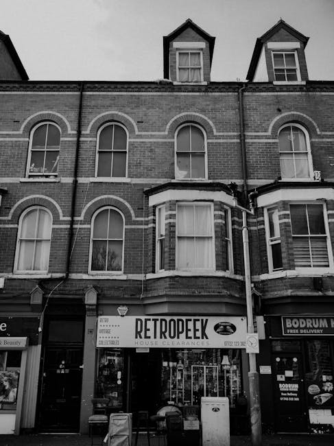 The image depicts the façade of a three-story brick building with arched and rectangular windows, some with visible curtains and window frames. The building’s exterior features a combination of red and brown brickwork with decorative white accents outlining the windows and edges. A large commercial sign reading 'RETROPEEK' advertising house clearances is mounted above the ground-floor shopfront, which includes a glass entrance door and display windows filled with various items. To the right of the site, another storefront offering food delivery services is partially visible. The ground level appears busy with the presence of small chairs and a waste bin near the entrance, suggesting an active commercial area. Above, the building's upper floors have small, dormer-style windows with steeply pitched roofs. The lighting is natural and consistent with daytime illumination, and the overall scene presents an urban street scene typical of property clearance or rubbish removal services operating in a busy town or city environment, with a focus on private waste handling outside local authority collection zones. The setting aligns with independent rubbish removal or house clearance activities, supporting the context of alternative waste management solutions offered by companies like House Clearance Tufnell Park.