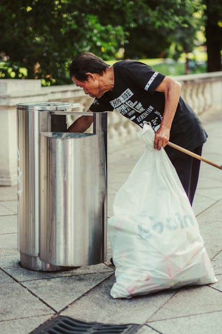 A middle-aged man with short dark hair, wearing a black T-shirt with white graphics, is seen disposing of waste into a cylindrical metal litter bin with a lid opening on top, located on a paved outdoor area. He is holding a white plastic bag filled with rubbish in his left hand and is leaning slightly forward while reaching into the bin with his right hand. The bin is positioned near a low, curved stone railing, and behind him, there is lush green foliage and trees, indicating a park or garden setting. The natural daylight highlights the reflective surface of the metal bin and the textures of his clothing and the ground. This scene relates to private waste collection or rubbish removal services often provided by companies like House Clearance Tufnell Park, emphasizing on-site or independent waste disposal efforts, especially in public outdoor spaces, supporting borough-specific rubbish clearance timings and restrictions.