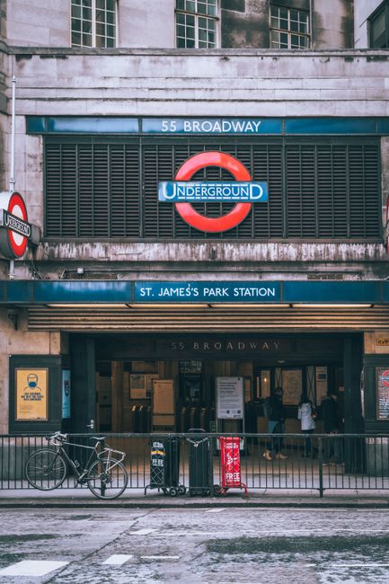 The image depicts the entrance to St. James's Park Station, part of the London Underground, situated beneath a building with a concrete facade. The station sign features a classic red and blue Underground roundel with the words 'UNDERGROUND' displayed prominently. Above the entrance, a white and blue sign indicates the station's location on 55 Broadway. The area in front of the station includes a sidewalk with a lead railing, a bicycle leaning against the railing, and a small red and white public information sign. The scene is set during daylight, with natural lighting illuminating the concrete and signage, and a few people visible inside the station entrance. The environment appears urban and well-maintained, with elements consistent with typical city infrastructure, highlighting its role as a hub for public transportation. This visual context indirectly relates to rubbish removal services by illustrating the city environment where waste management might be necessary for surrounding commercial and residential areas, reflecting the need for independent waste disposal or private rubbish collection. House Clearance Tufnell Park, specializing in rubbish services, operates within such urban settings, supporting efficient waste handling in busy city environments.