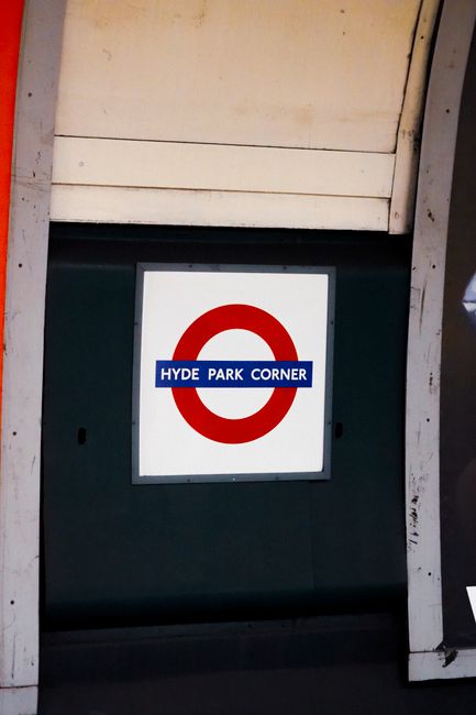 A close-up view of a square sign mounted on a dark green wall or panel, positioned beneath a beige or light-colored overhang or structural element. The sign features a red circular symbol, similar to a London Underground roundel, with a blue rectangular band across the center displaying the white text 'HYDE PARK CORNER.' The surrounding environment appears to be part of a public or commercial space, possibly at a bus or subway station, with visible metal framing and signage support structures. The area is well-lit, with no visible debris or rubbish in the immediate vicinity, suggesting the scene is clean and maintained. This signage exemplifies typical public transport or directional signs in an urban setting, aligning with the context of nearby rubbish removal or public service facilities often adjacent to such infrastructure.