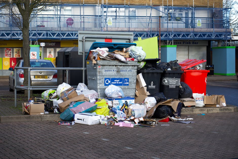 In the foreground of the image, there is a large collection of overflowing rubbish bins and scattered waste on a paved outdoor area. The waste includes various cardboard boxes, plastic bags, paper packaging, and miscellaneous household refuse, with some items spilling onto the pavement. The bins are of different colours, including a large central grey bin marked for mixed paper and cardboard, and smaller black and red bins beside it. The grey bin’s lid is partially open, revealing paper and cardboard waste inside. Surrounding the bins are piling-up bags of garbage, some black and others white, along with loose items such as plastic containers and packaging. To the left, a grey vehicle is parked behind a metal railing, with a few smaller boxes and waste items placed against the railing on the pavement. In the background, there is a closed shopfront with their signage visible, and a building under scaffolding with a blue safety netting, indicating ongoing construction or maintenance work. The overall scene depicts an area awaiting rubbish collection, illustrating the need for efficient waste removal services, such as those provided by House Clearance Tufnell Park.